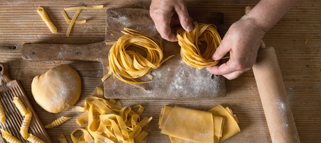 Zelf pasta maken: handen draaien verse tagliatelle tot nestjes, naast pastadeeg, garganelli en een houten deegroller